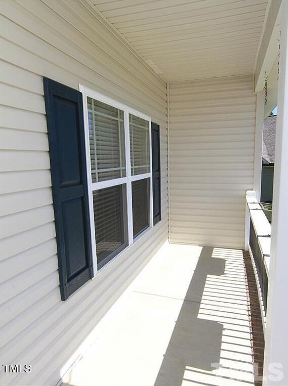 A narrow porch with beige siding and a white railing, featuring a window with dark blue shutters. The porch casts striped shadows from the sunlight filtering through the railing.