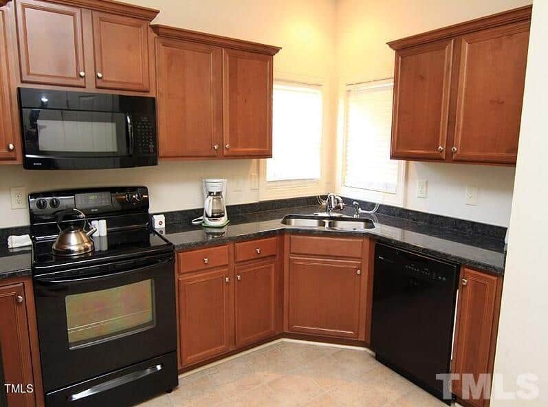A kitchen with wooden cabinets, a black stove, microwave, and dishwasher. A kettle sits on the stove, and a coffee maker is on the counter. Two windows provide natural light above the corner sink. The floor is tiled.