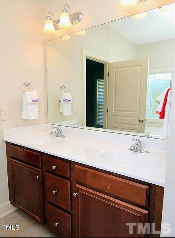 Modern bathroom interior with a large mirror above a double sink vanity. The vanity has a wooden base and white countertop. Towels hang on rings beside the mirror, and warm lighting illuminates the space. A partially open door is visible in the background.