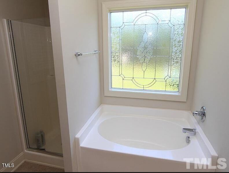 A bathroom featuring a white bathtub below a decorative glass window with a floral pattern. Next to the bathtub is a glass-enclosed shower. The walls are painted white, creating a clean and bright atmosphere.