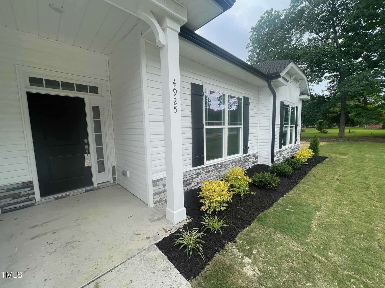 A white house with black shutters, a covered front entry, and a landscaped garden bed with yellow and green shrubs alongside a green lawn and tall trees in the background. House number 4925 is visible on a pillar.