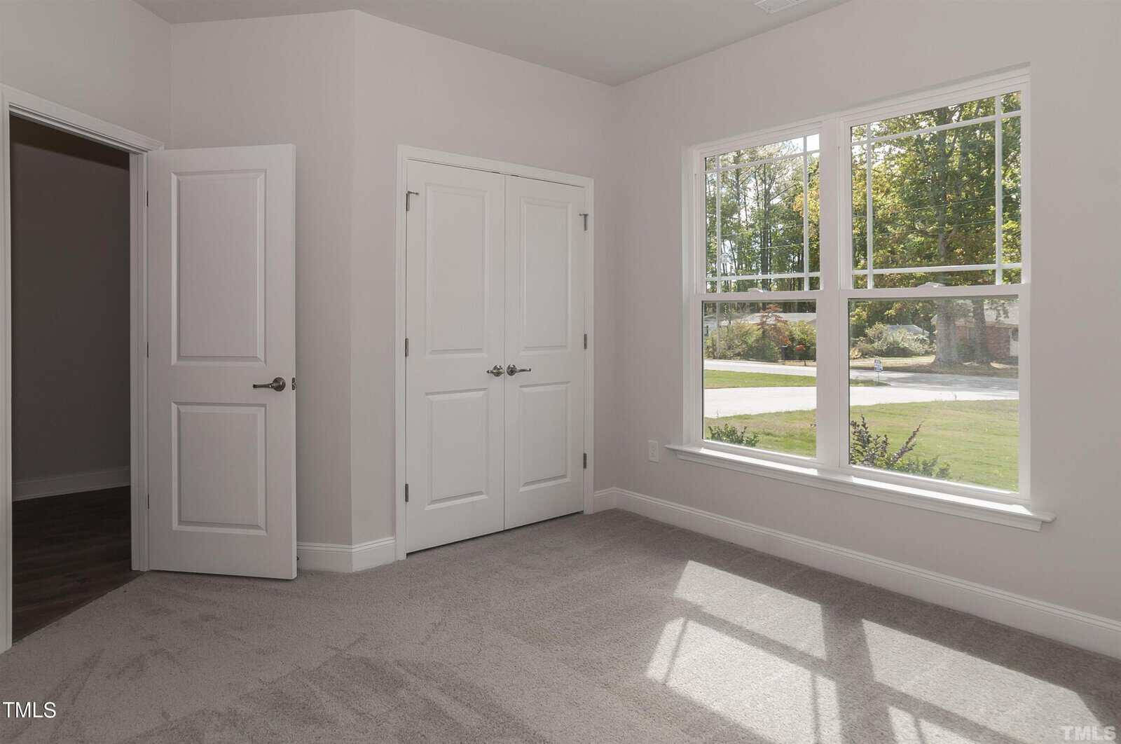 A bright, empty room with light beige walls, carpeted floor, a double-door closet, and a large window letting in natural light, showing a view of greenery outside.