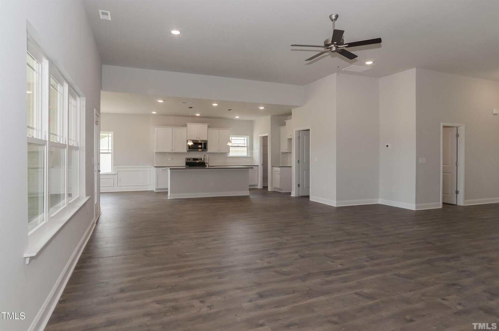Spacious modern open-concept living area with large windows, light gray walls, dark wood flooring, and a ceiling fan; kitchen with white cabinets and island visible in the background.