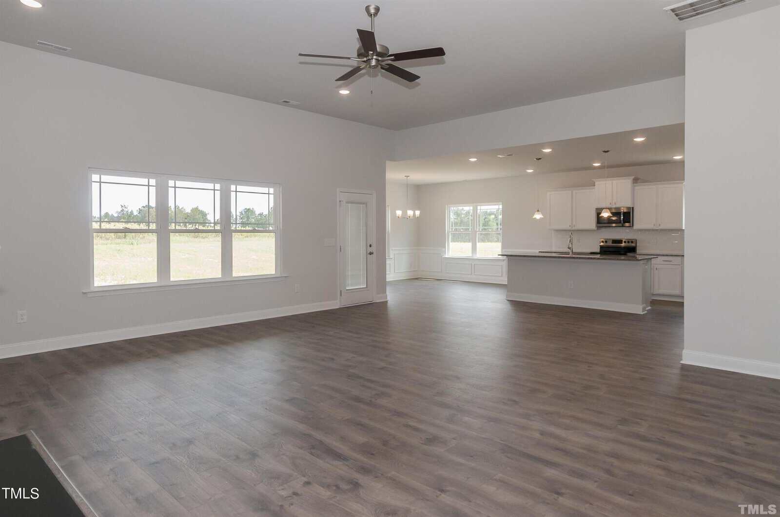Spacious, empty open-concept living area with wood flooring, large windows, ceiling fan, and a view into a modern white kitchen and dining space with pendant lights.
