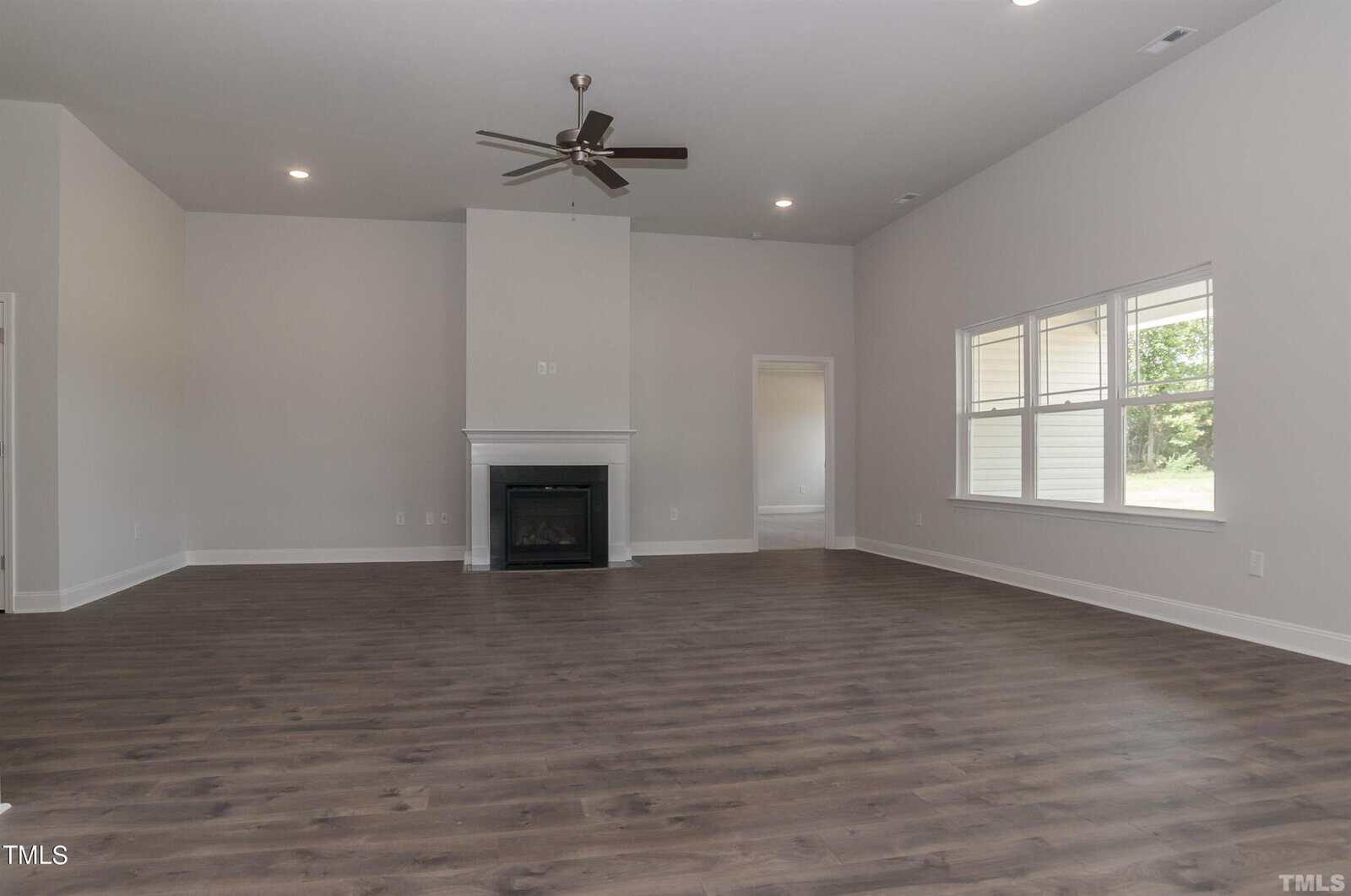 Spacious empty living room with light gray walls, dark wood floors, a ceiling fan, fireplace, and large windows letting in natural light. A doorway leads to another room.