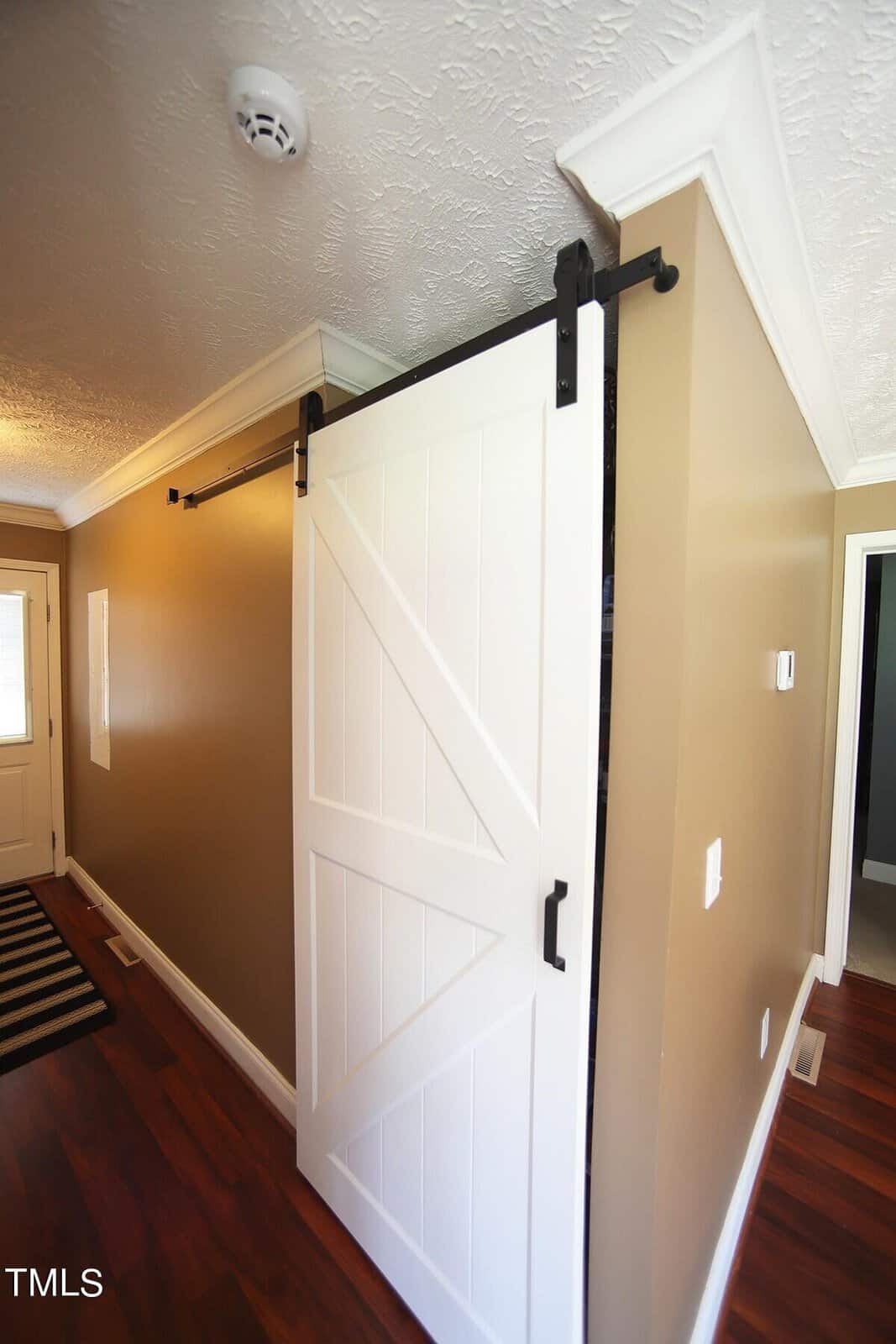 A hallway with beige walls and white crown molding features a white sliding barn door with black hardware. The door is partially open, revealing another room. The floor is wood, and a door mat is present at the entrance on the left. A smoke detector is on the ceiling.
