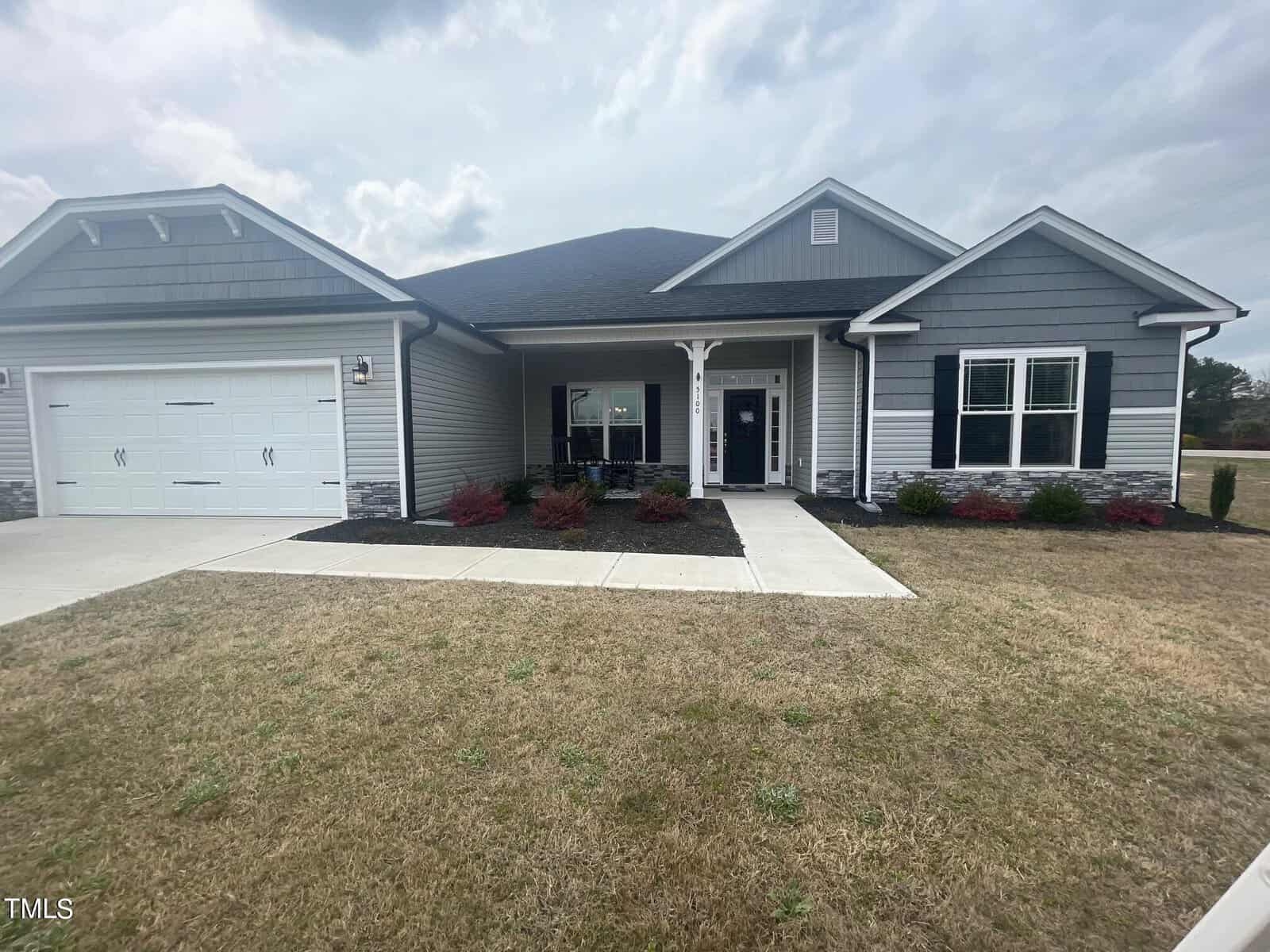 A single-story gray house with white trim, a covered front porch, double garage doors, and a well-maintained lawn with shrubs and a concrete walkway leading to the entrance under a cloudy sky.