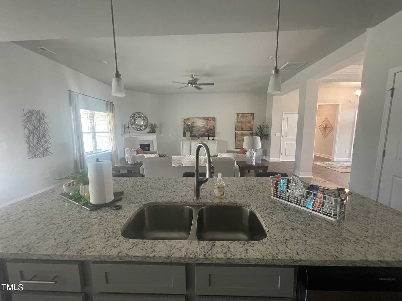 View from a granite kitchen island with a double sink, looking into a bright living room with sofas, a coffee table, decor, and a fireplace. Large windows and pendant lights illuminate the open space.