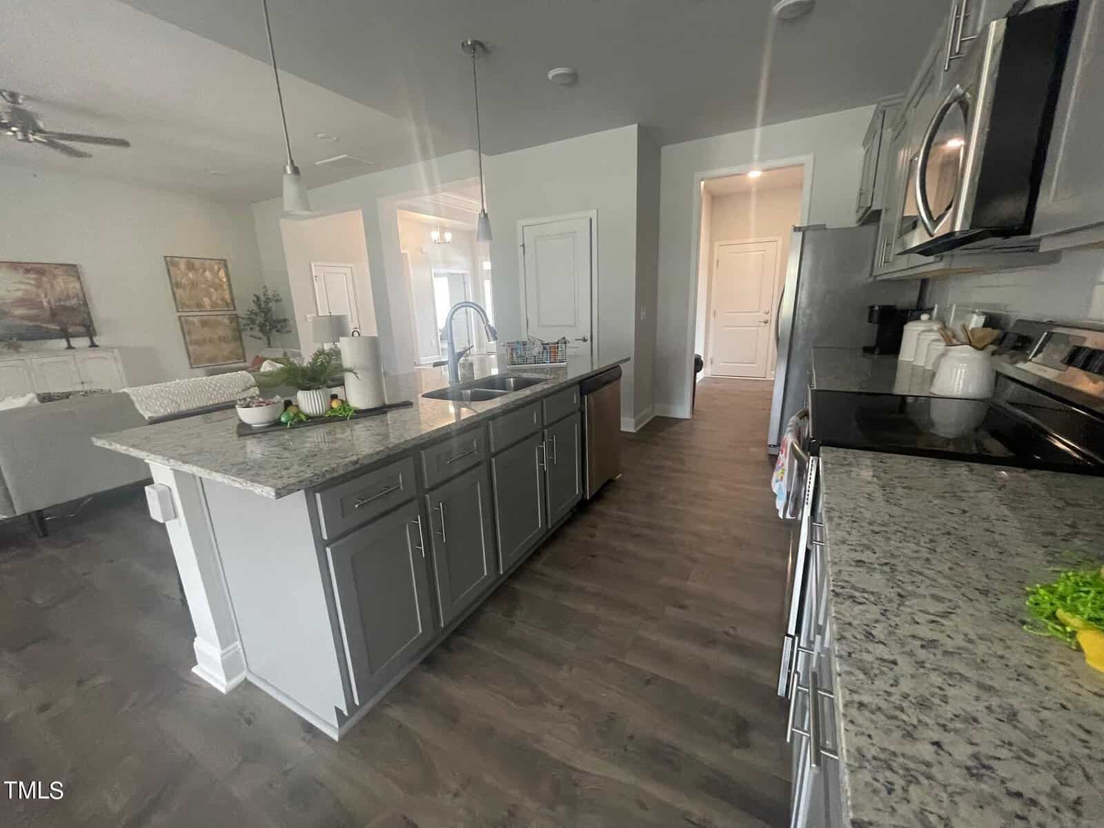 A modern kitchen with gray cabinets, granite countertops, a large island with a sink, stainless steel appliances, and hardwood floors. The kitchen opens to a bright living and dining area in the background.