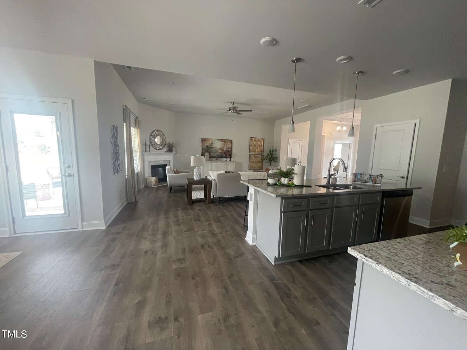 A spacious, modern open-concept living area with wood-look flooring, a kitchen island with barstools, granite countertops, and a cozy living room with white furniture and a fireplace in the background. Natural light fills the space.