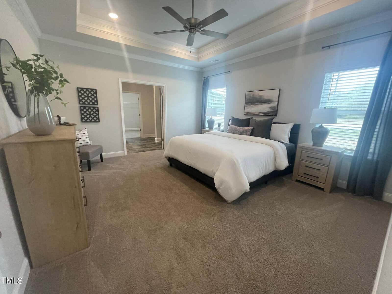Spacious bedroom with a large bed, nightstands, and lamps by two windows. There's a ceiling fan, dresser with a plant, wall art, and neutral-colored walls and carpet. Double doors lead to an adjacent bathroom.