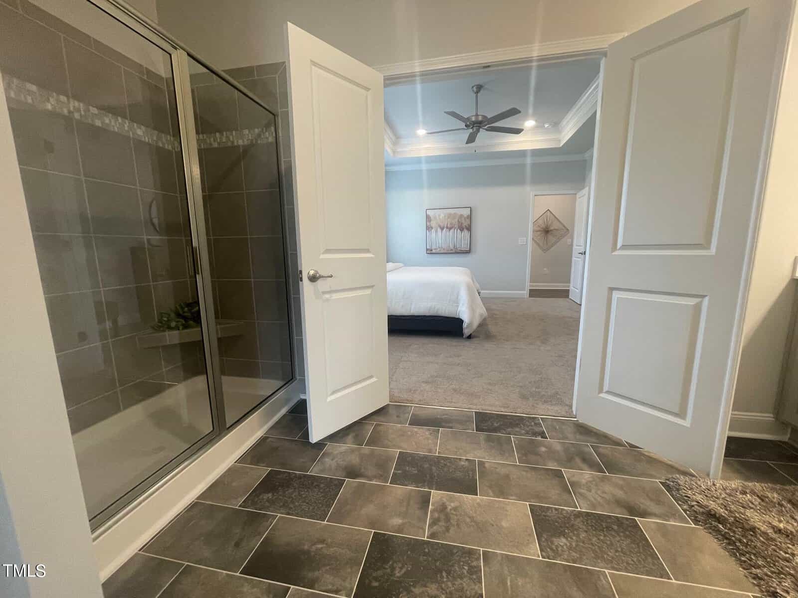 A view from a bathroom with tiled floors and a glass shower, looking into a carpeted bedroom with a bed, ceiling fan, and wall art through open double doors.