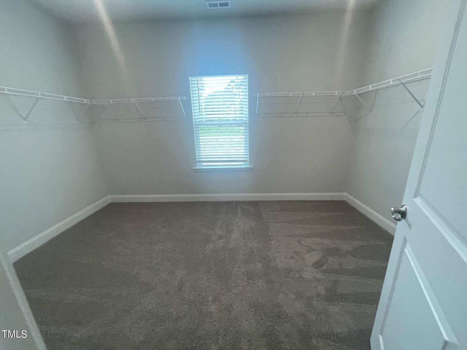 Empty walk-in closet with light gray walls, carpeted floor, white wire shelving along three walls, a window with blinds centered on the far wall, and an open white door.