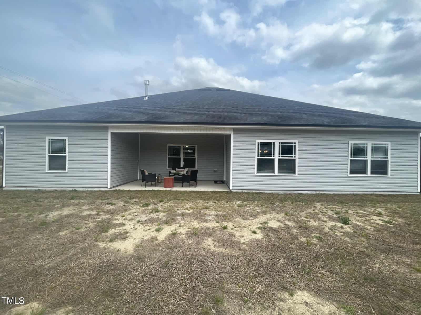 A single-story house with gray siding, a dark roof, and three windows on the back. There is a covered patio area with outdoor furniture and a fire pit, and a patchy lawn in the foreground under a cloudy sky.