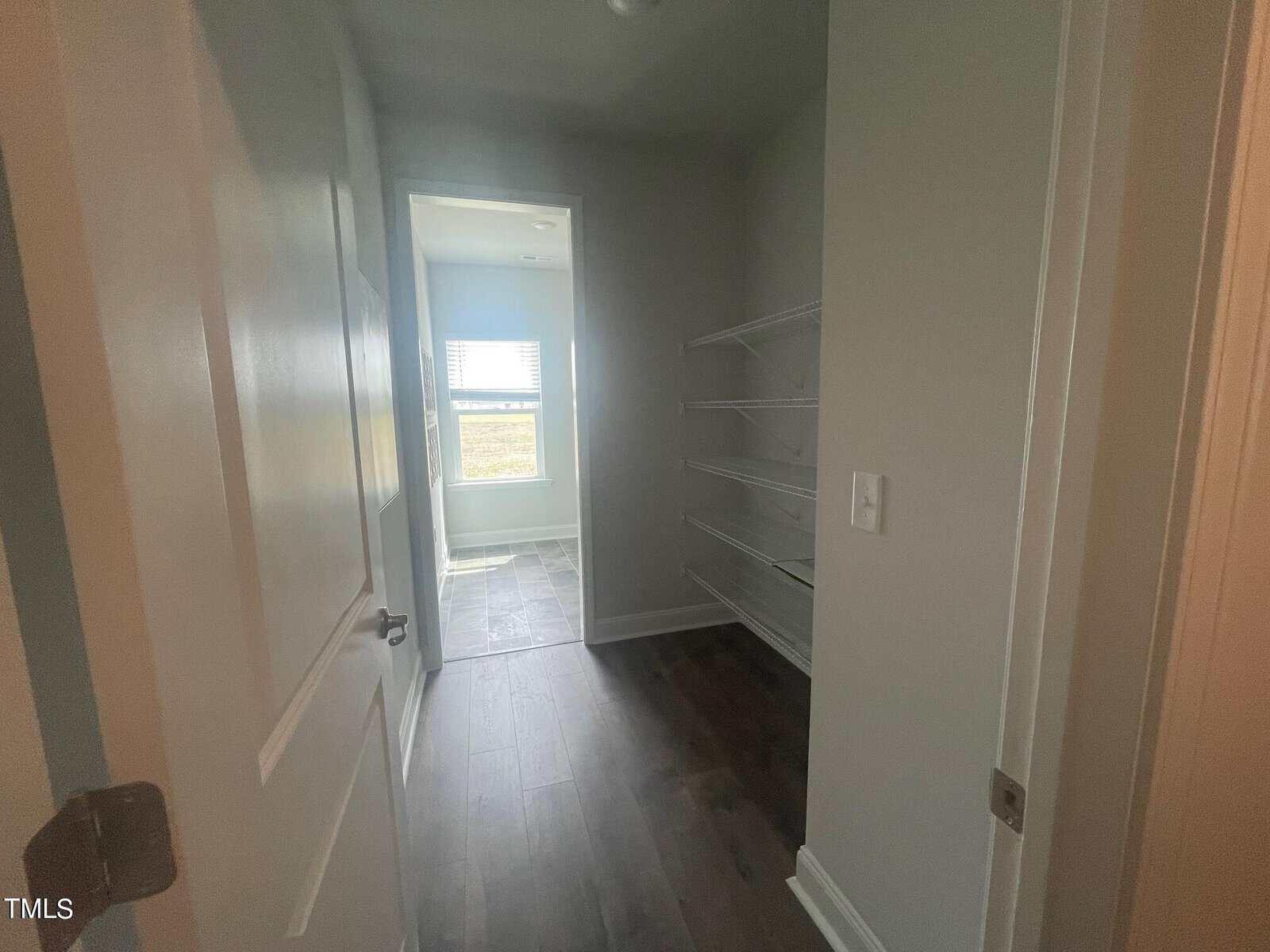 A small walk-in closet with several white wire shelves on the right wall, leading to a bright bathroom with a window at the far end. The floor has dark wood and gray tile.