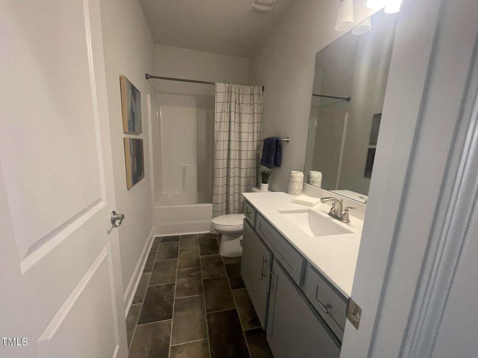 Modern bathroom with gray floor tiles, white vanity with two sinks, a large mirror, and a shower-tub combo with a white plaid curtain. Two framed artworks hang on the left wall above the toilet.