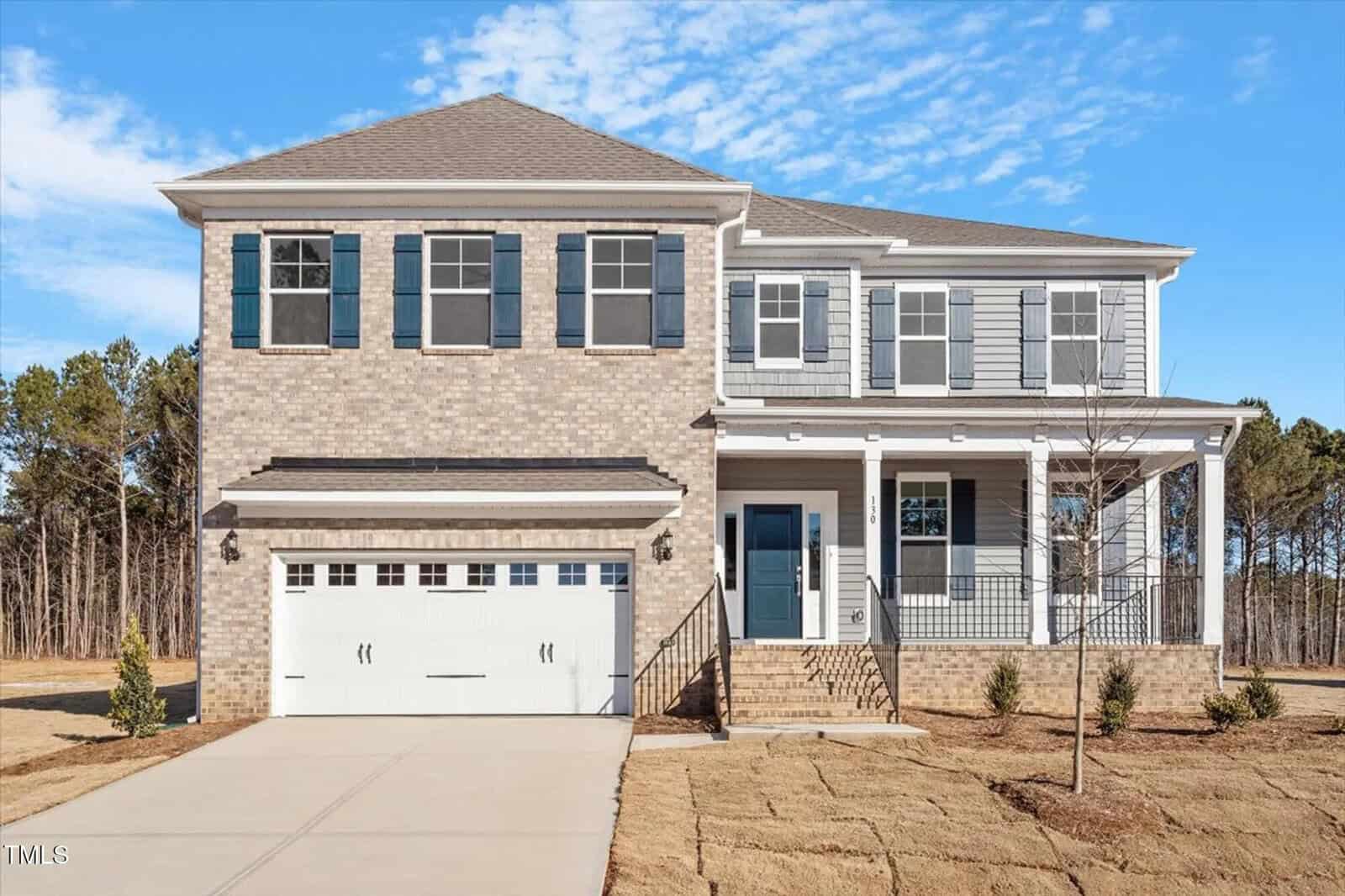 Two-story modern suburban house with brick and siding exterior, blue shutters, double garage, and front porch. The home sits on a new lawn with a backdrop of trees and a clear sky.