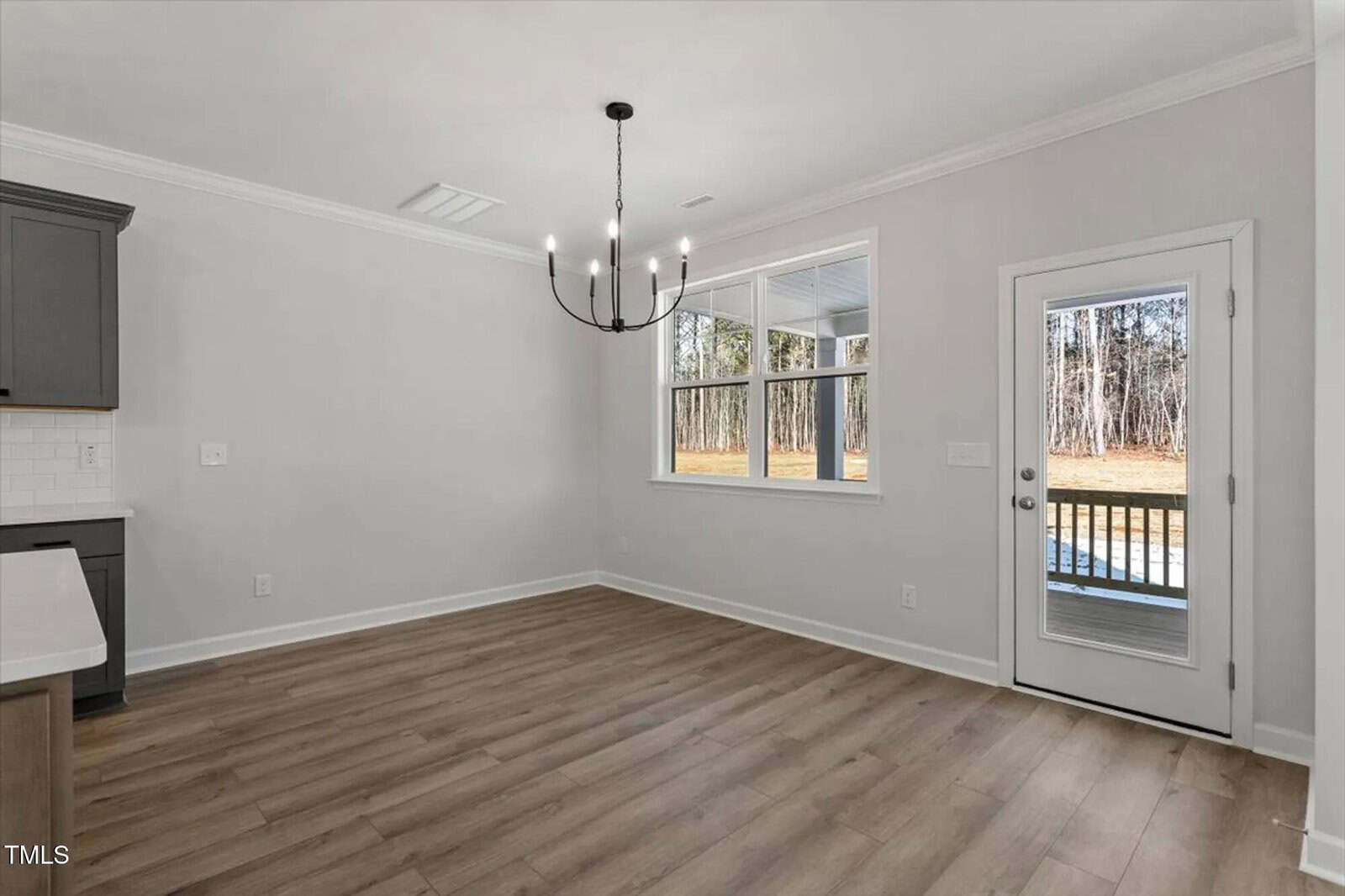 Bright dining area with light wood floors, a modern black chandelier, large window, and glass door opening to a deck with a view of trees and grass outside. Light gray walls and white trim create a fresh, open feel.