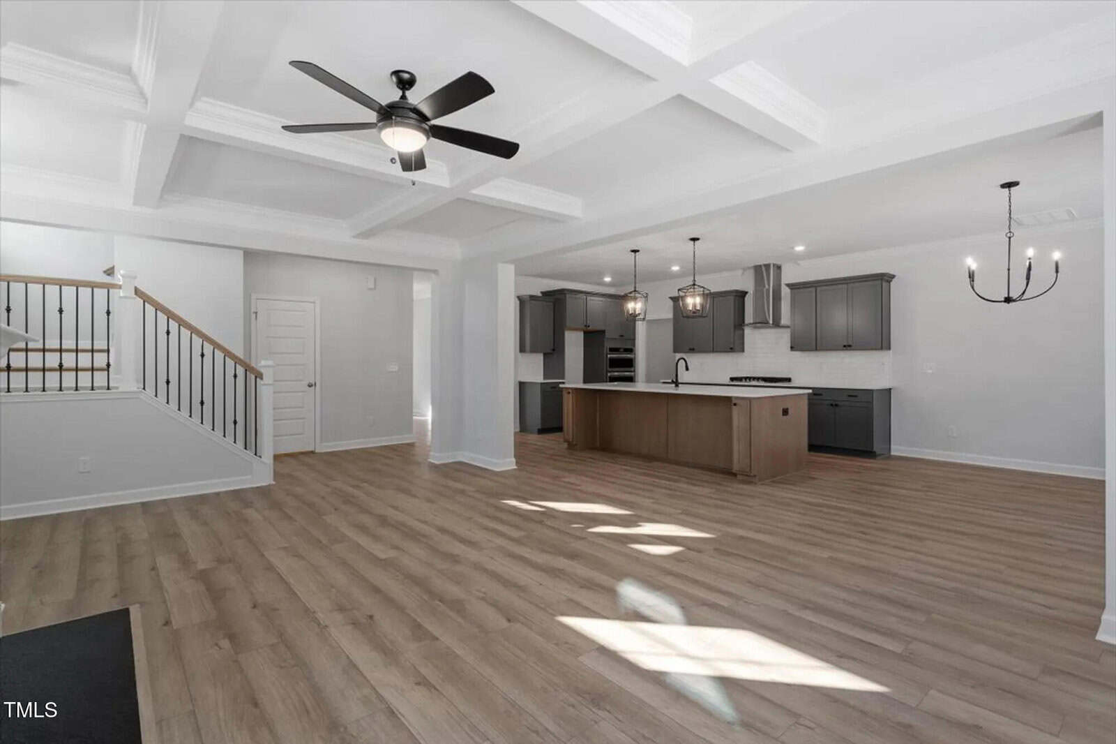 Open-concept living area with light wood flooring, a coffered ceiling with a ceiling fan, a modern kitchen with a large island, gray cabinets, and two black chandeliers. Staircase visible on the left.