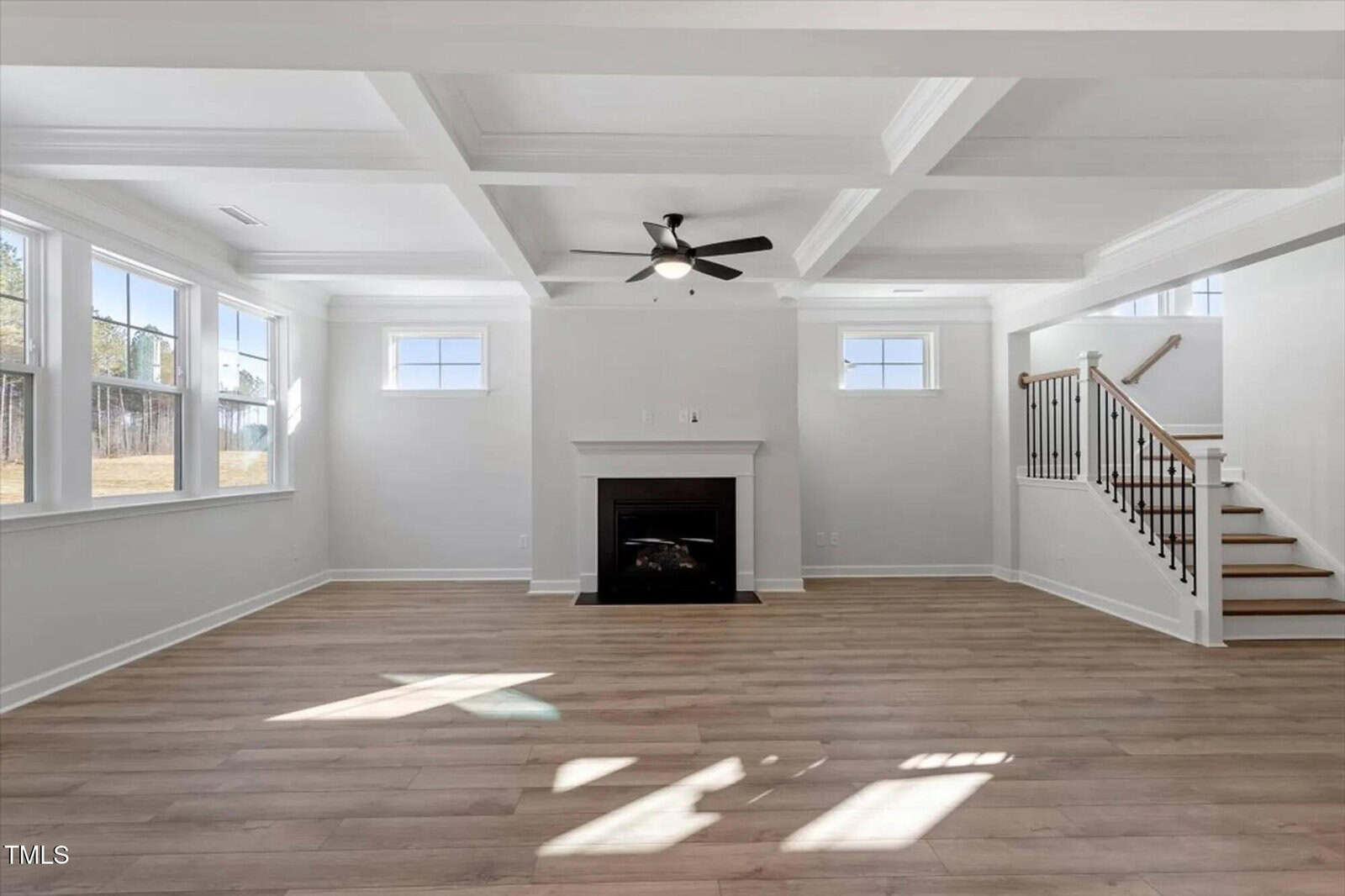 Bright, spacious living room with large windows, a ceiling fan, coffered ceiling, a central fireplace, light wood floors, and a staircase with wooden handrails on the right.