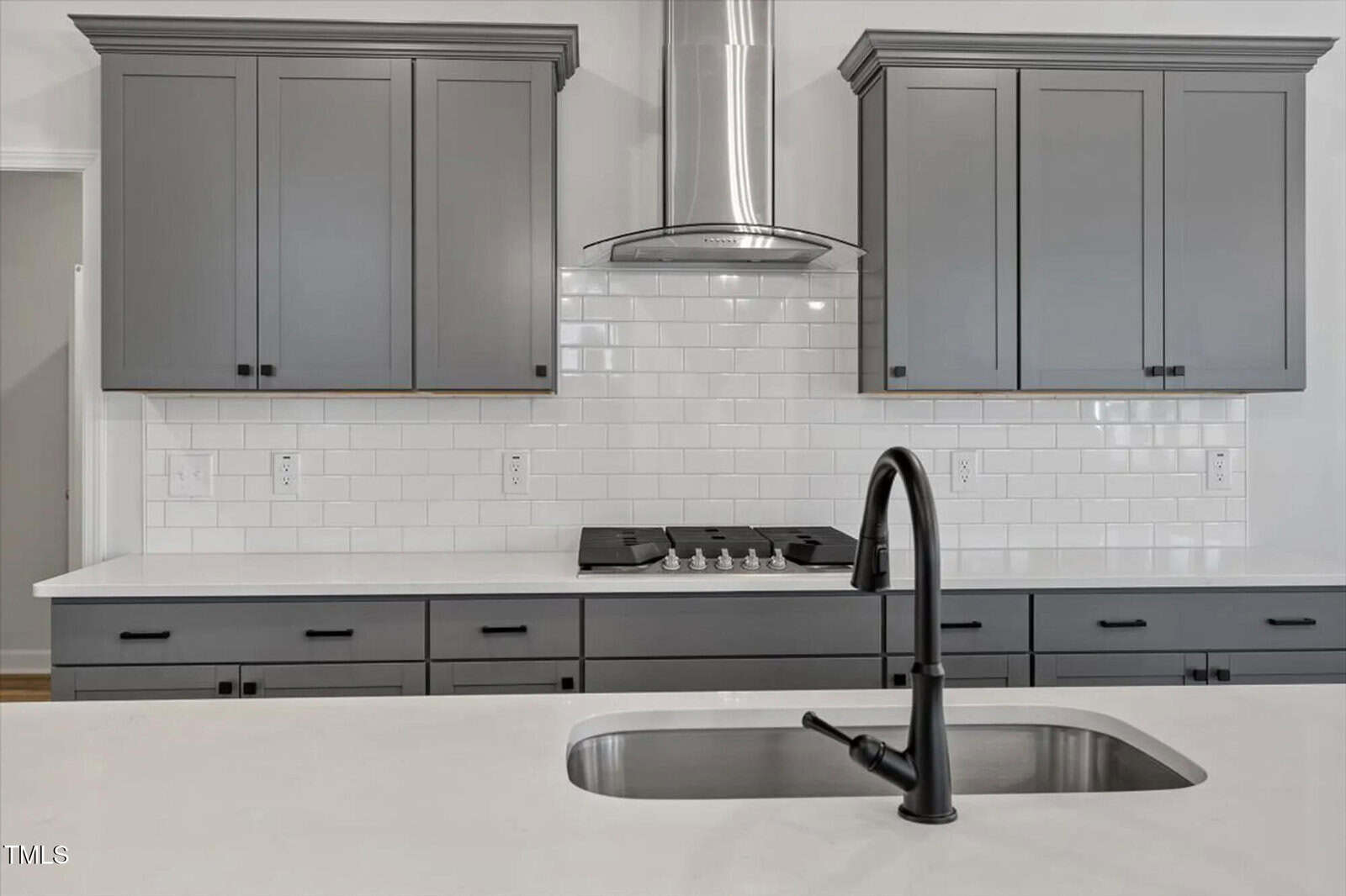 Modern kitchen with gray cabinets, white subway tile backsplash, stainless steel range hood, gas cooktop, and a white countertop island with an undermount sink and black faucet in the foreground.