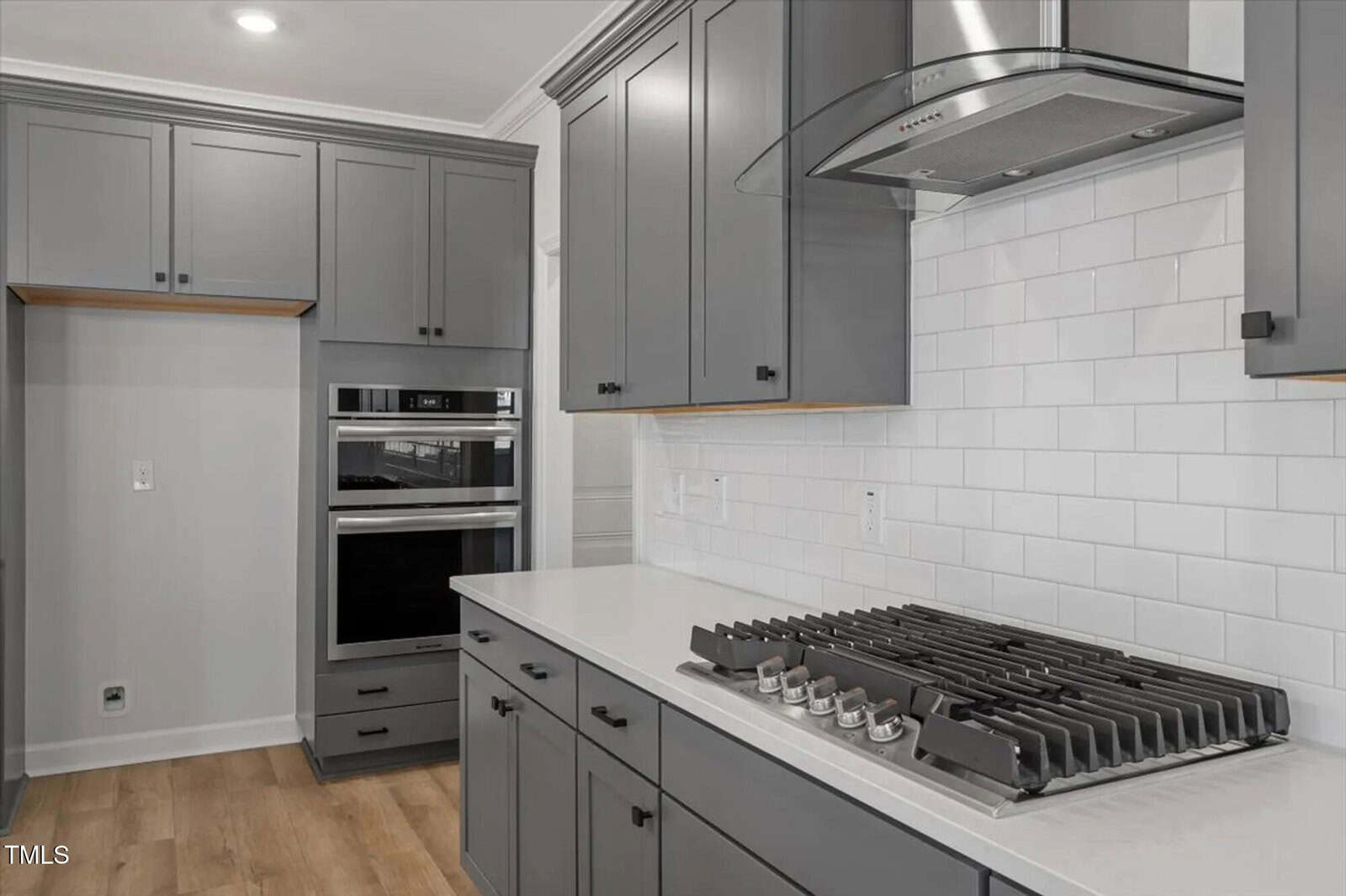 Modern kitchen with gray cabinets, white subway tile backsplash, stainless steel gas cooktop, built-in double oven, wood flooring, and under-cabinet lighting.