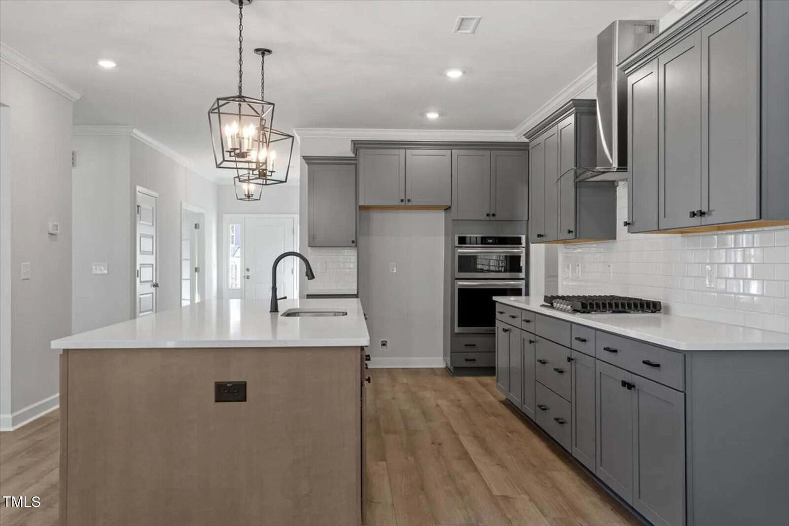 Modern kitchen with gray cabinets, white subway tile backsplash, a large island with a sink, stainless steel appliances, wood flooring, and two geometric pendant lights above the island.