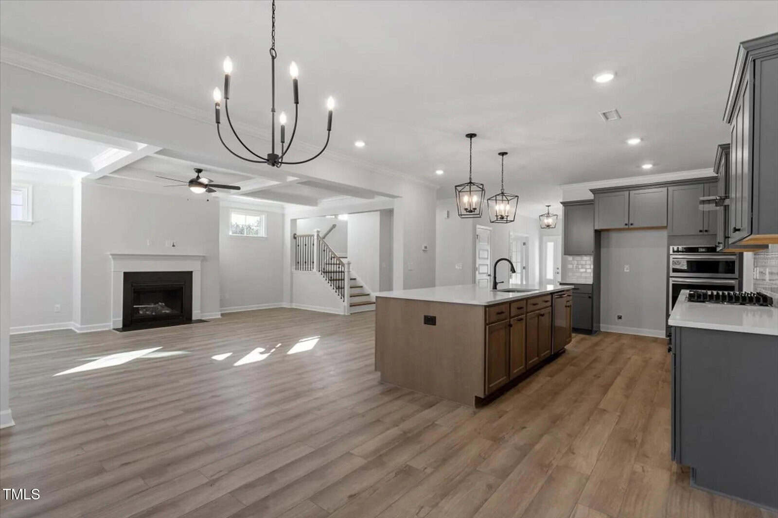 Spacious modern kitchen and living area with light wood floors, gray cabinets, a large island with pendant lights, a chandelier, fireplace, and a staircase leading upstairs. Bright natural light fills the open space.
