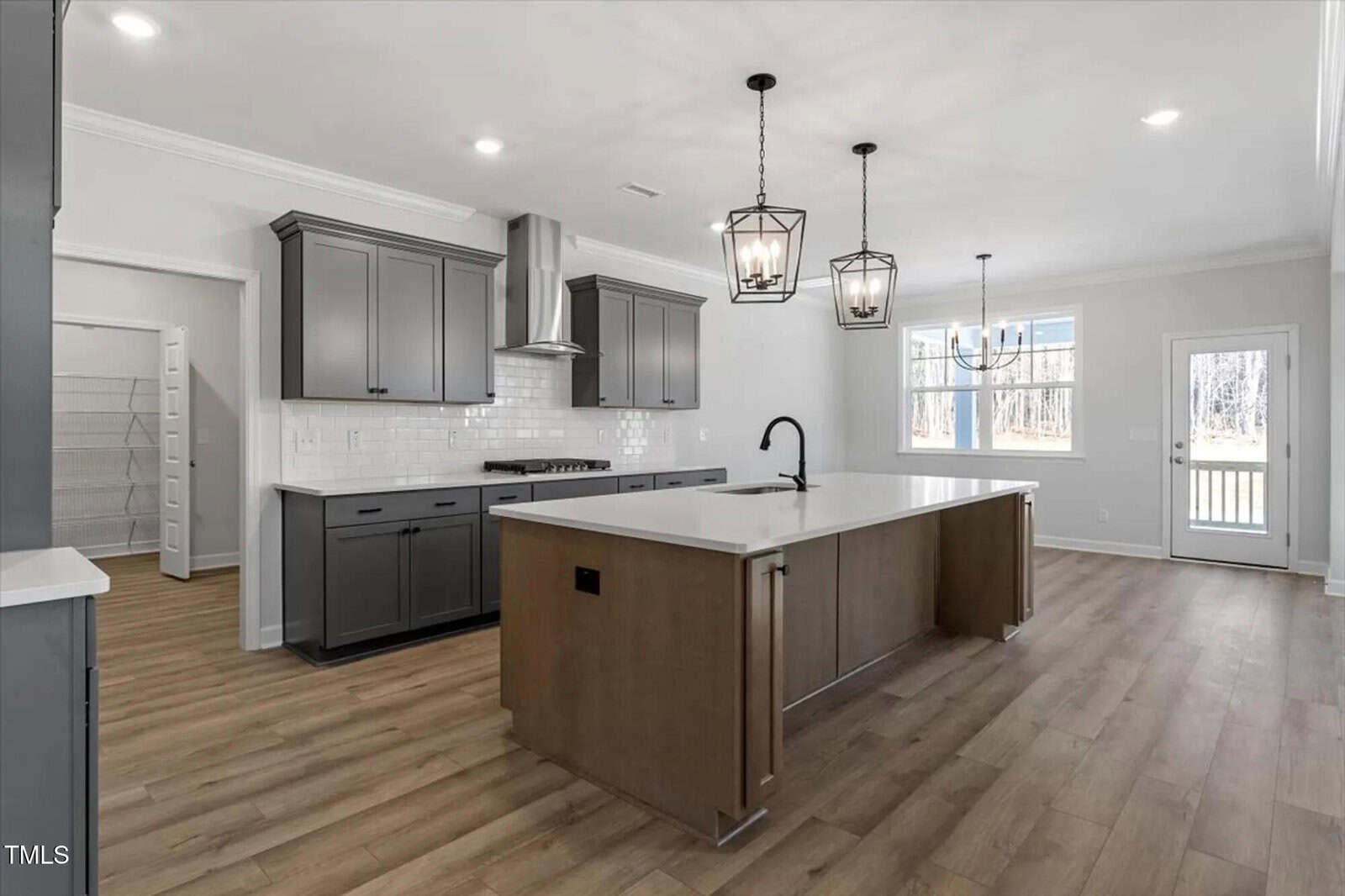 Modern kitchen with gray cabinets, white countertops, stainless steel appliances, a large center island with a sink, pendant lights above, wood-style flooring, a pantry, and a window and door leading outside.