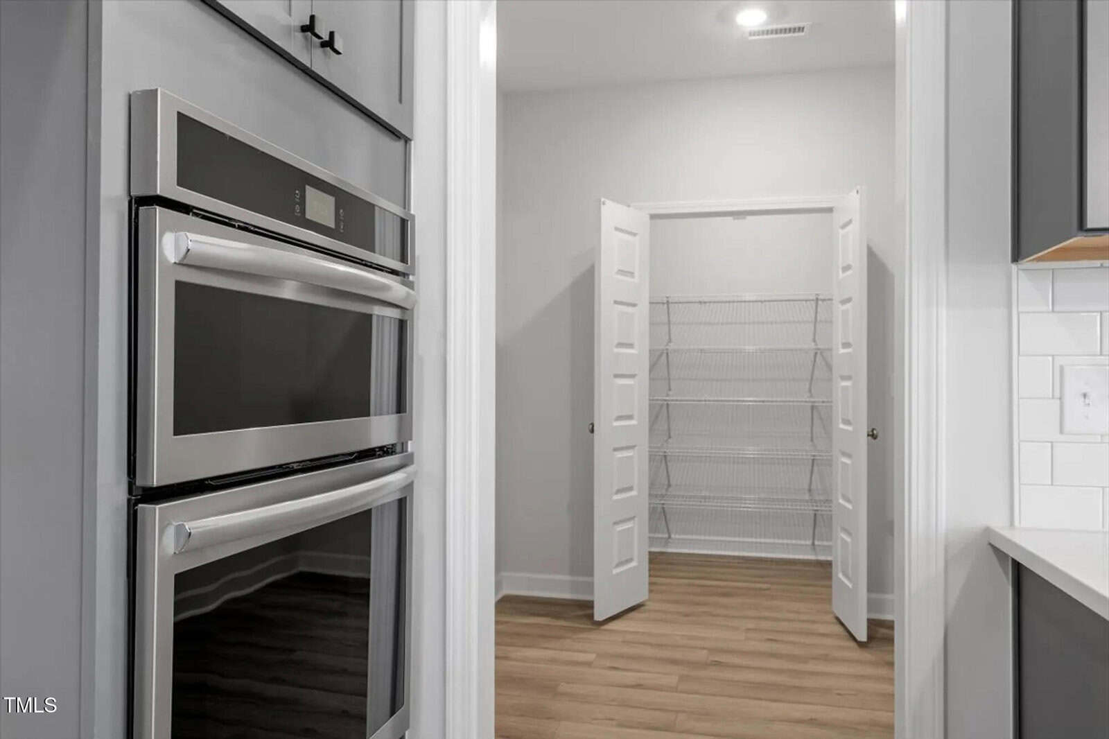 A modern kitchen with built-in stainless steel double ovens on the left and an open pantry with white double doors and wire shelving in the background. The floor is light wood.