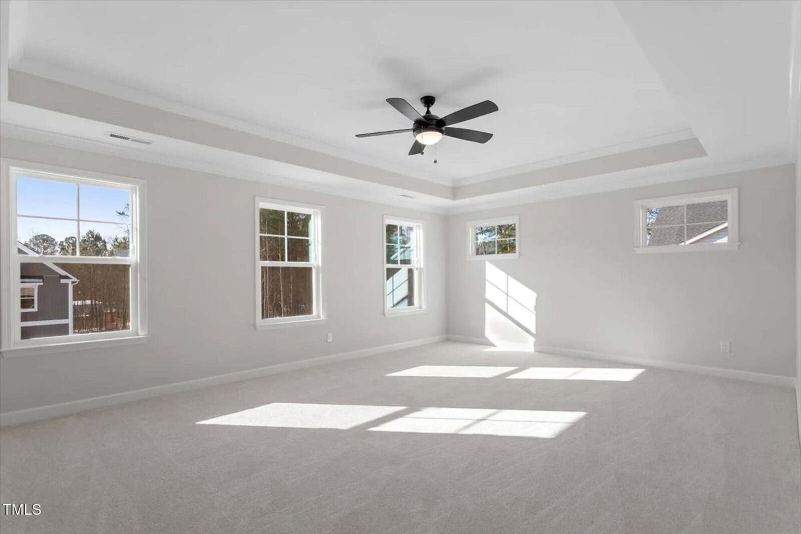 Spacious, empty bedroom with light gray walls, white trim, carpeted floor, five windows, and a black ceiling fan in the center of a tray ceiling. Sunlight streams through the windows onto the floor.