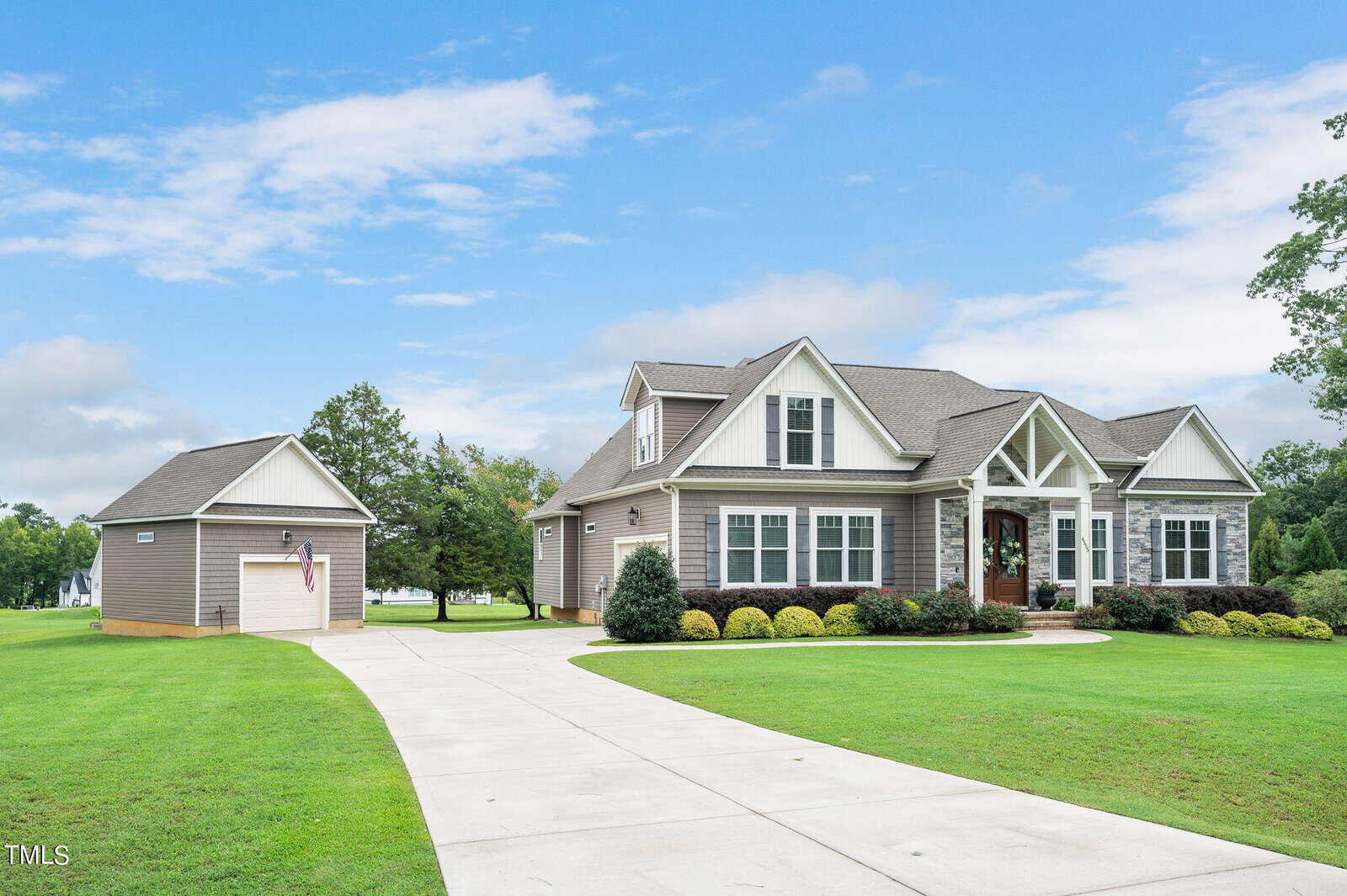 A large suburban house with beige siding, white trim, and a gray roof, surrounded by a well-manicured lawn. There's a separate garage to the left and a long driveway lined with greenery under a partly cloudy sky.