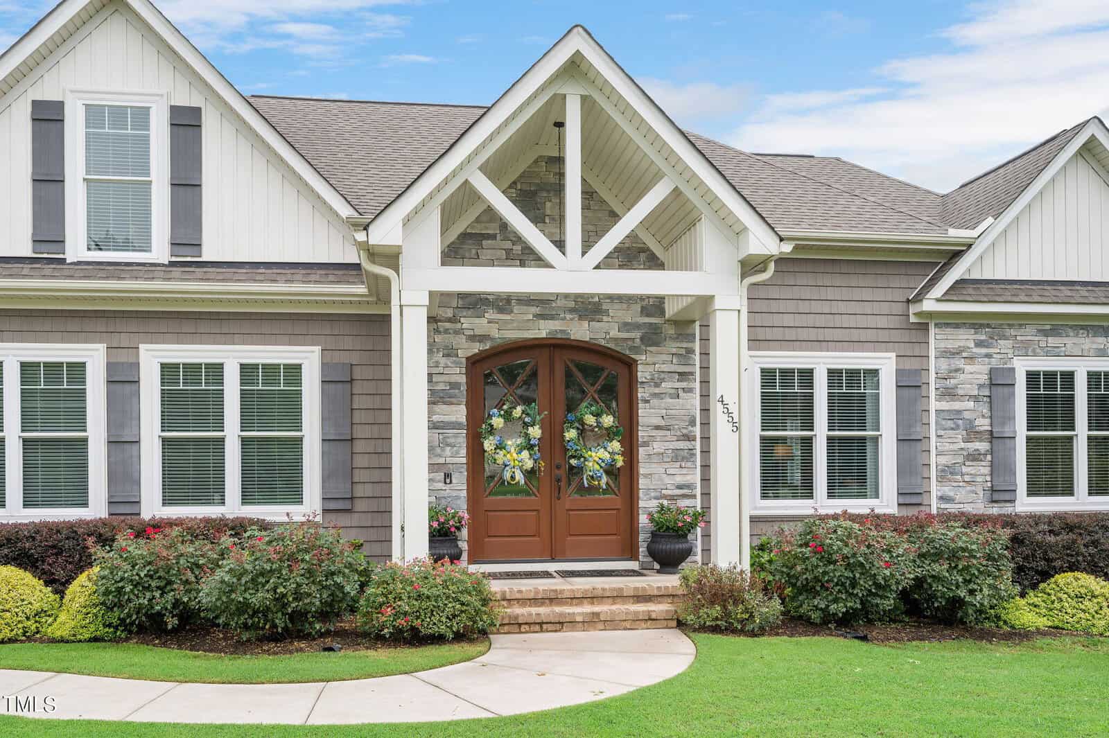 Front view of a modern house with stone and siding exterior, featuring large wooden double doors and decorative wreaths. The entrance is flanked by windows with white shutters, and the yard has manicured bushes and a curved pathway.