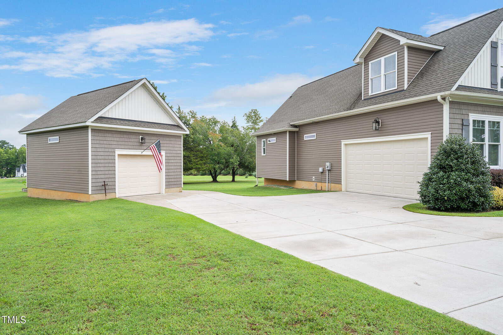 A suburban home with gray siding, a large driveway, and a neatly trimmed lawn. An American flag is mounted on a smaller adjacent shed. The sky is clear and blue, providing a calm atmosphere.