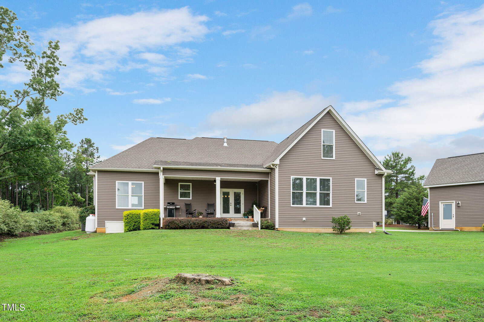 A single-story house with a gray exterior and a small front porch is set in a grassy yard. The sky is partly cloudy. A garage is visible on the right, with an American flag displayed. Trees are in the background.