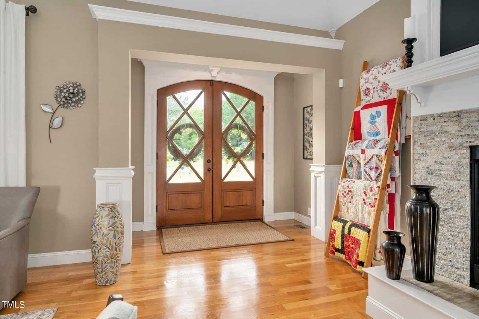 A welcoming foyer with wooden double doors featuring glass panels. The space has light-colored walls, a vase on the floor, and quilts on a ladder rack next to a stone fireplace. Natural light brightens the hardwood floor.