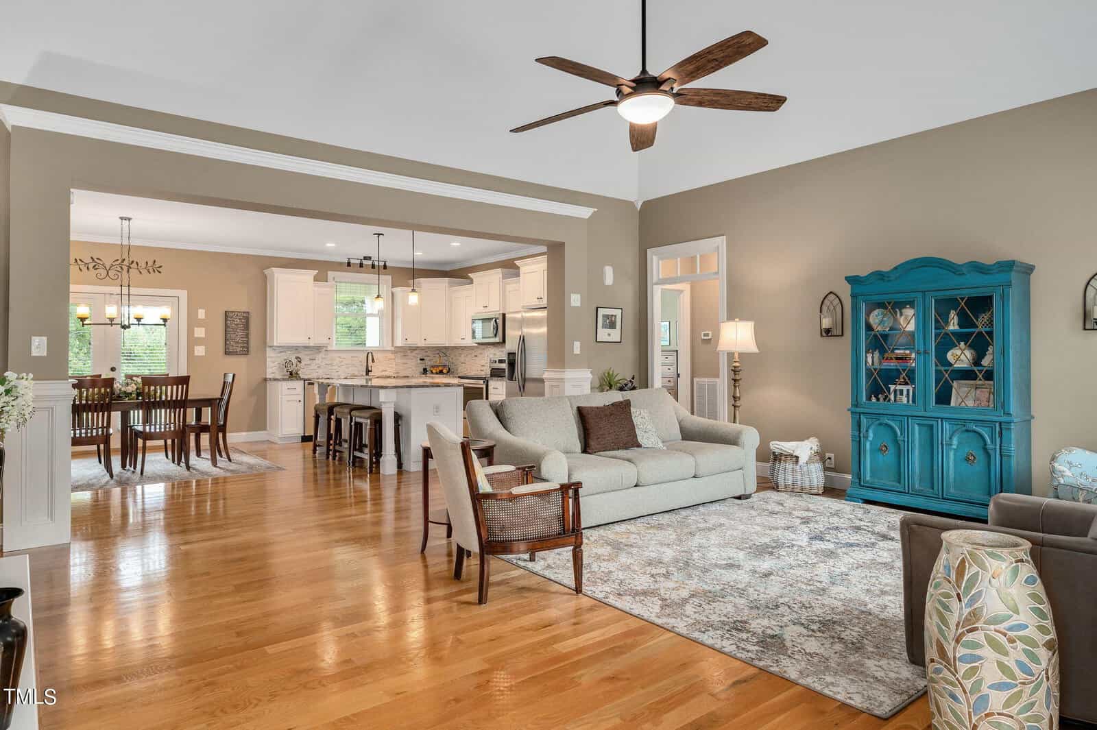 Open concept living area with a gray sofa, patterned rug, and ceiling fan. A turquoise cabinet stands against a brown wall. The kitchen features white cabinetry and a breakfast bar. A dining table is visible in the adjacent room. Wood flooring throughout.