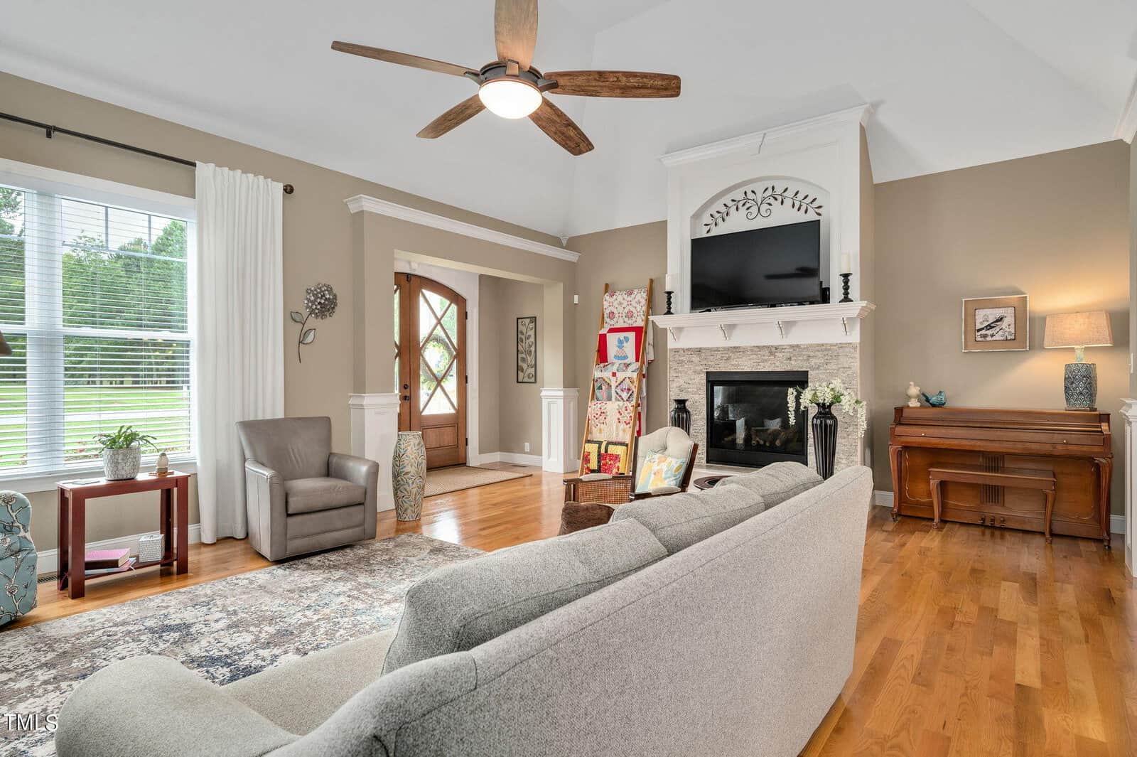 A cozy living room with a gray sectional sofa facing a fireplace and TV. The room features a ceiling fan, a piano, and a large window with curtains. A patterned rug, an armchair, and decorative plants are present. A wooden door is in the background.