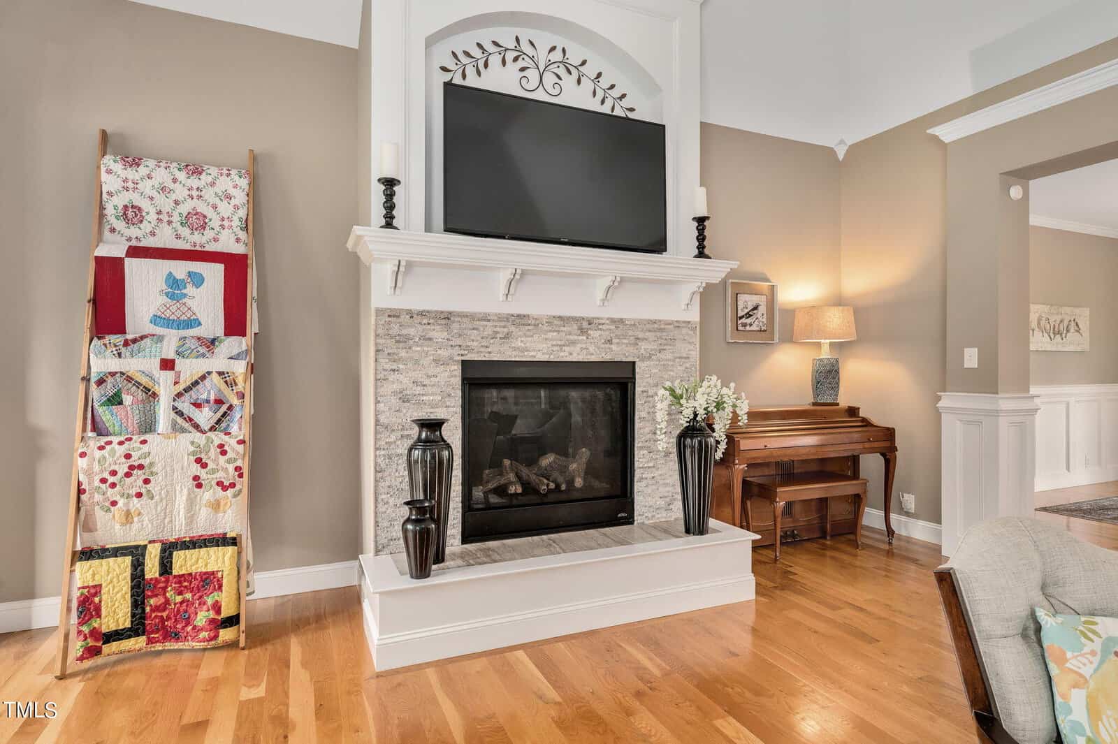 A cozy living room with a stone fireplace and a TV mounted above it. A quilt ladder displays colorful quilts to the left, while a wooden piano sits in the corner. A lamp and floral arrangement add warm touches to the room's decor.
