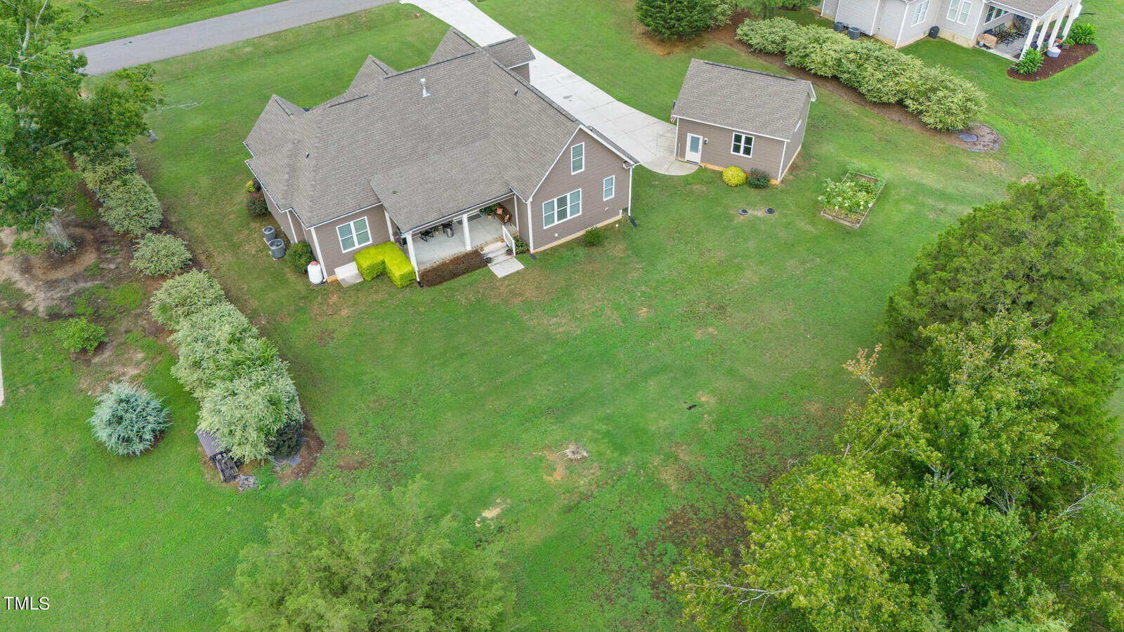 Aerial view of a suburban house with a brown roof, surrounded by a large green lawn, trees, and shrubs. The driveway leads to a garage. Nearby, there are additional houses and well-maintained landscaping.