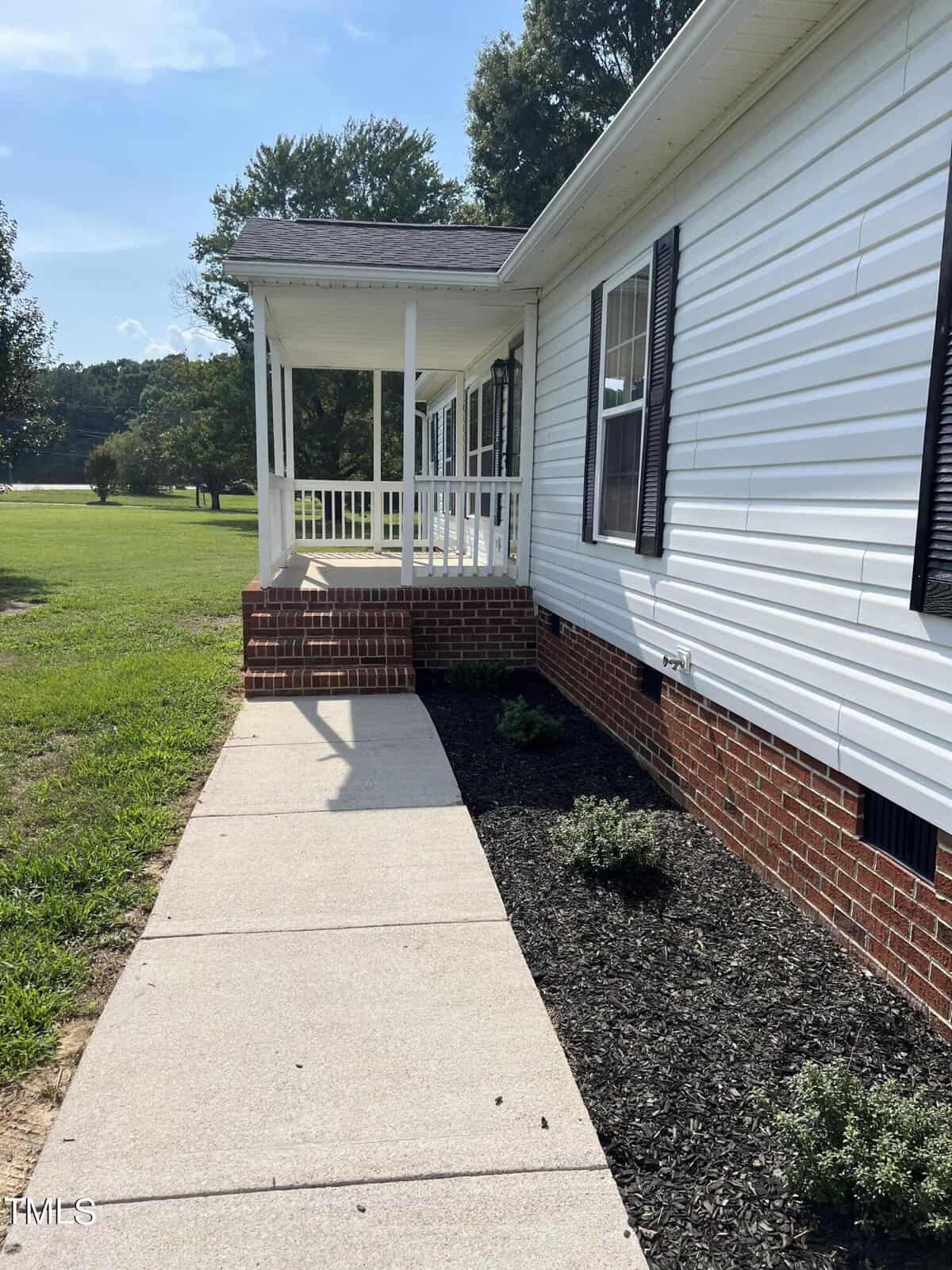A concrete pathway leads to the entrance of a white house with black shutters. The house has a small porch with white railings and brick steps. Mulched flower beds with small shrubs line the house's foundation. The yard is grassy with trees in the background.