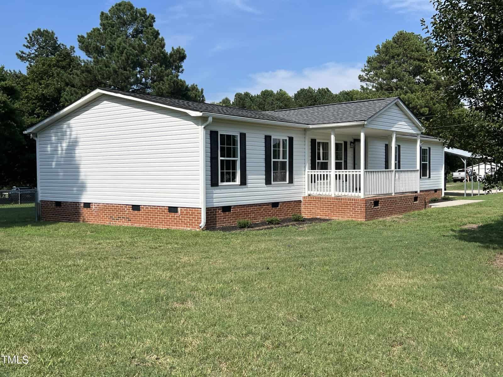 A white single-story house with black shutters, a brick foundation, and a small front porch is shown. The house is situated on a grassy lawn with trees in the background, and the sky is clear and blue.