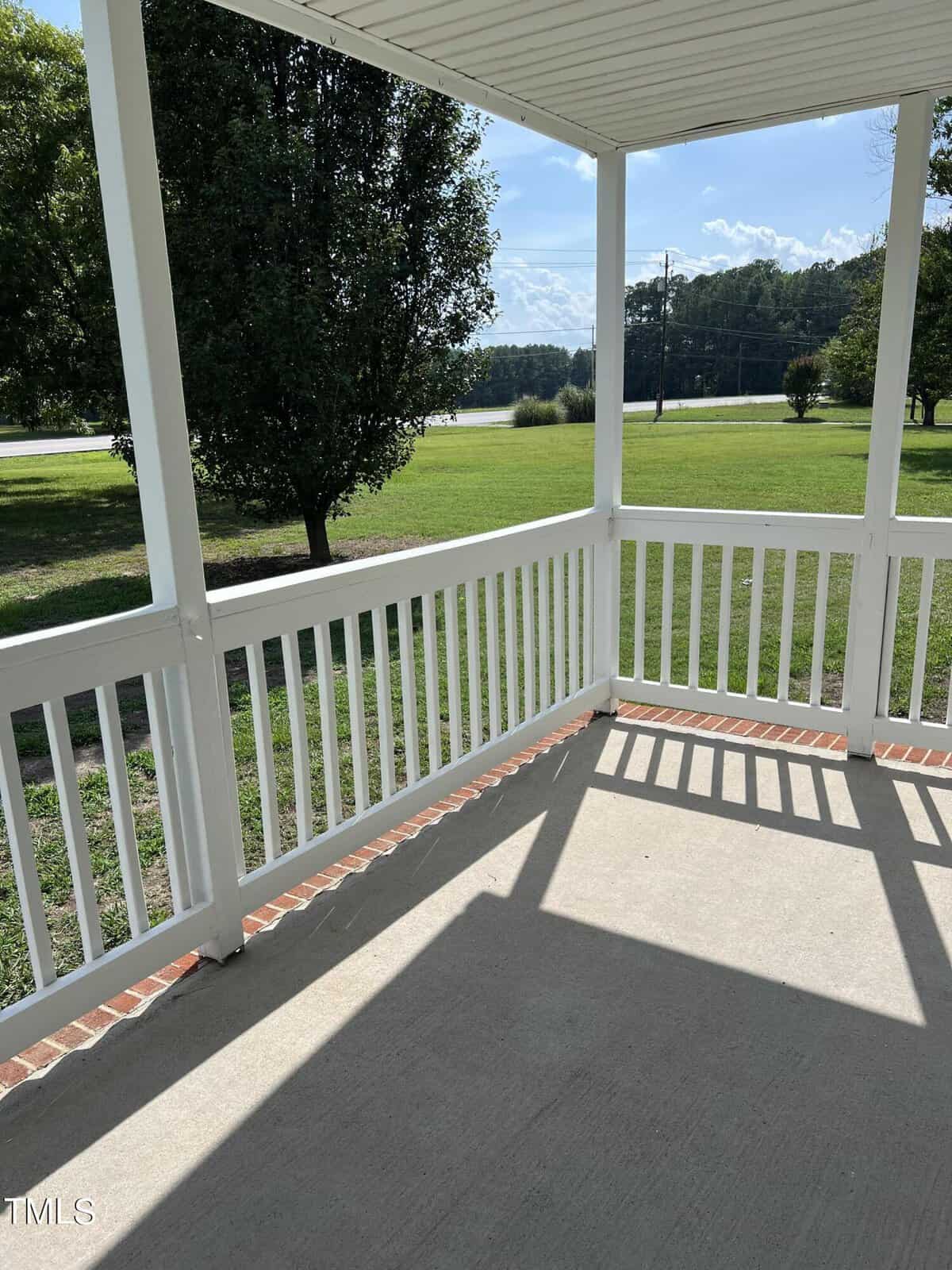 A covered porch with white railings and a concrete floor offers a view of a grassy lawn and trees in the background. Sunlight creates shadows on the porch, and a road is visible in the distance.