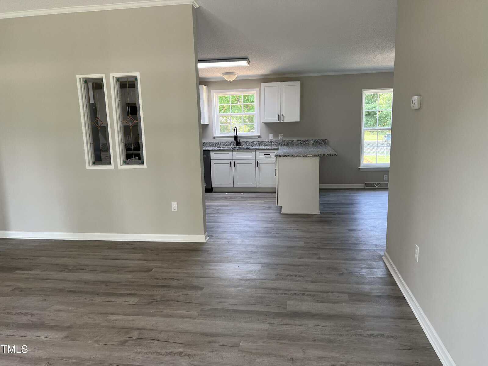 A modern, open-concept kitchen with light grey wooden flooring is shown. The kitchen features white cabinets, a granite countertop, a stainless steel sink, and large windows that let in ample natural light.