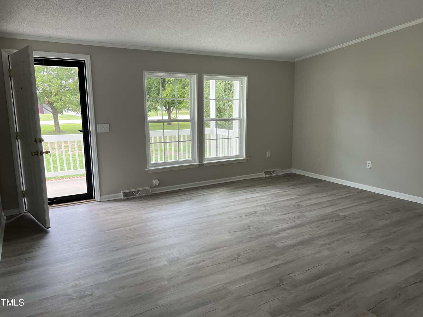 An empty living room with light gray walls, two large windows, and a door to the left that is slightly open. The floor is covered with light-colored laminate wood. Outside the windows, greenery and part of a porch railing are visible.