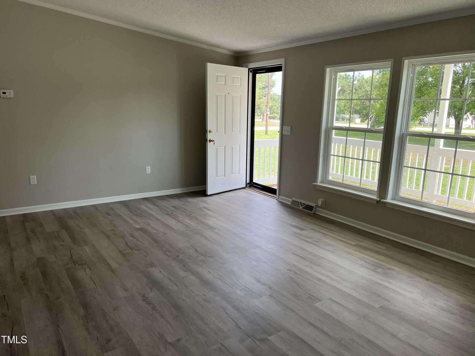 A spacious, empty living room with light gray walls and hardwood floors. The room features a white front door, partially open, and two large windows allowing natural light to fill the space. The windows offer a view of greenery outside.