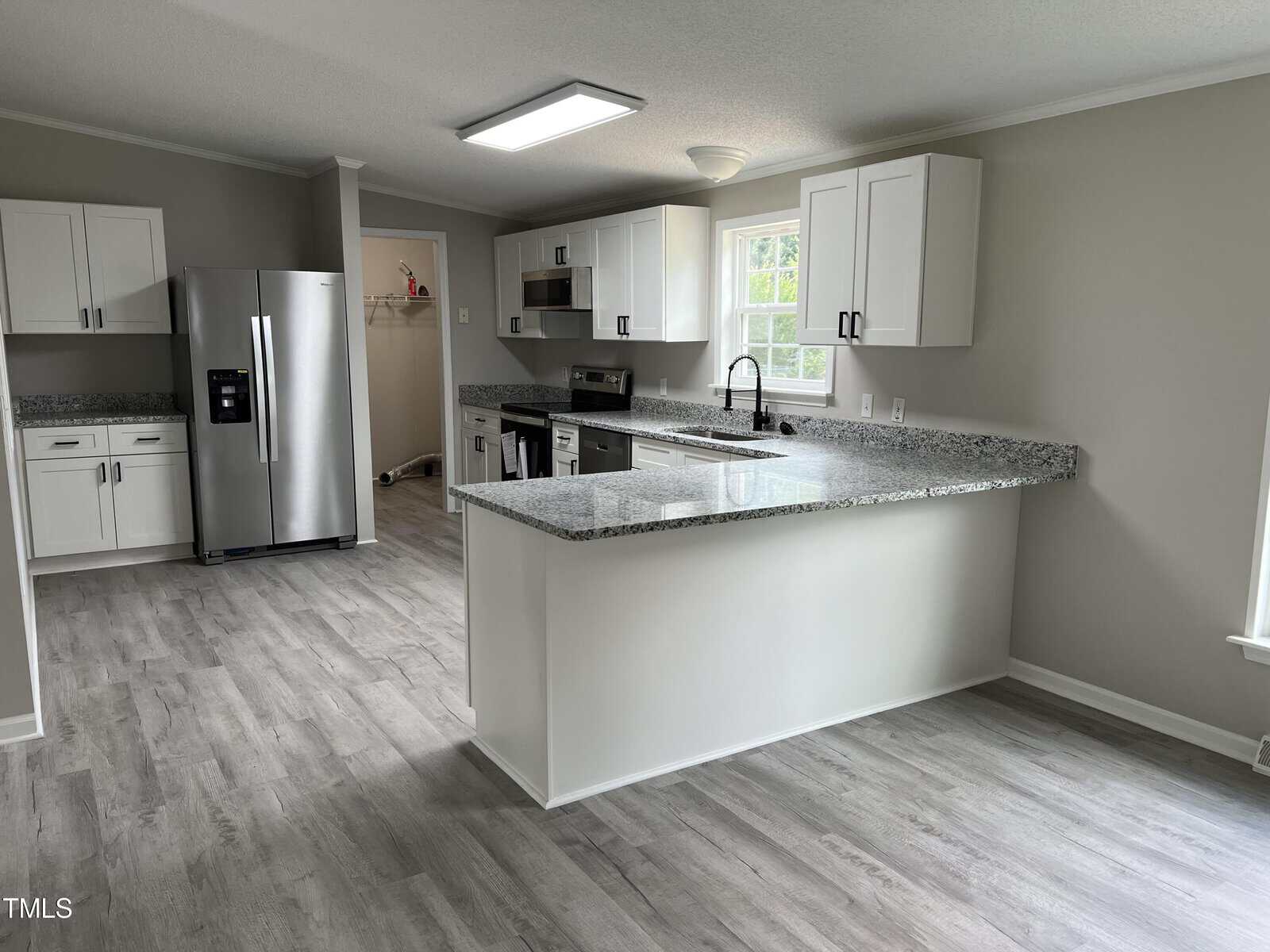 A modern kitchen features light gray wood-like vinyl flooring, white cabinets with black handles, a granite countertop, and stainless steel appliances including a refrigerator, oven, and microwave. There is a window above the sink and a laundry room visible in the background.