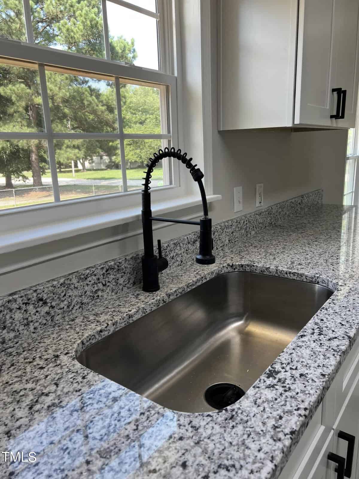 A kitchen sink with a high-arc black spring faucet set in a countertop made of speckled granite. The countertop extends along the wall below a large window, providing a view of a grassy yard with trees. White cabinets with black handles are seen on the right side.