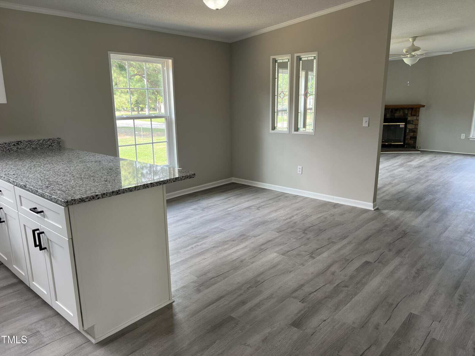 Open living area with light gray walls, large windows, wood-style flooring, white kitchen cabinets with granite countertops, and a view toward a cozy room with a fireplace and ceiling fan.