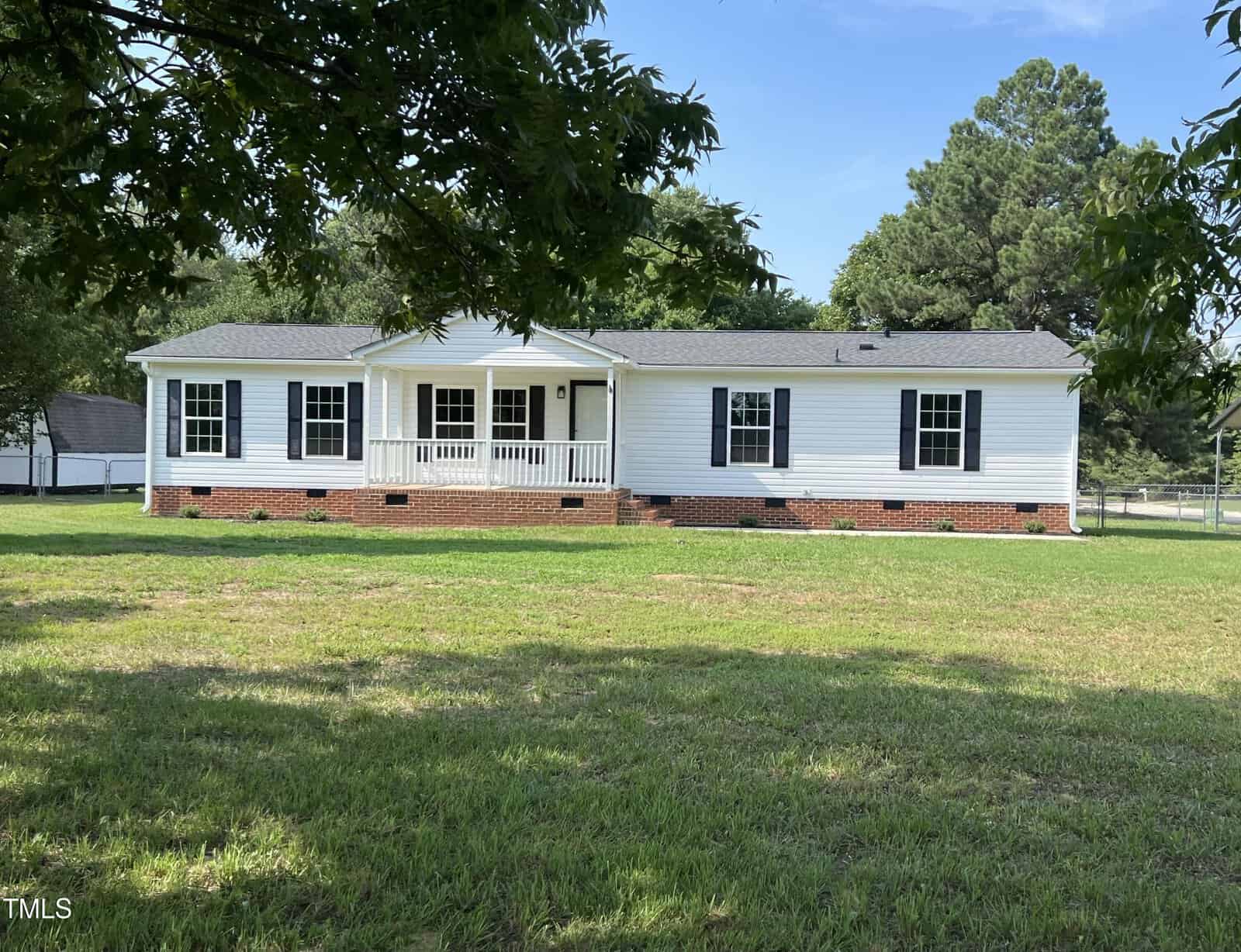 A single-story white house with black shutters at 35 Twelve Oaks Lane, Henderson, features a small covered front porch and a brick foundation. Set in an expansive grassy yard with trees partially in view and a fence in the background, the scene is framed by a clear sky.